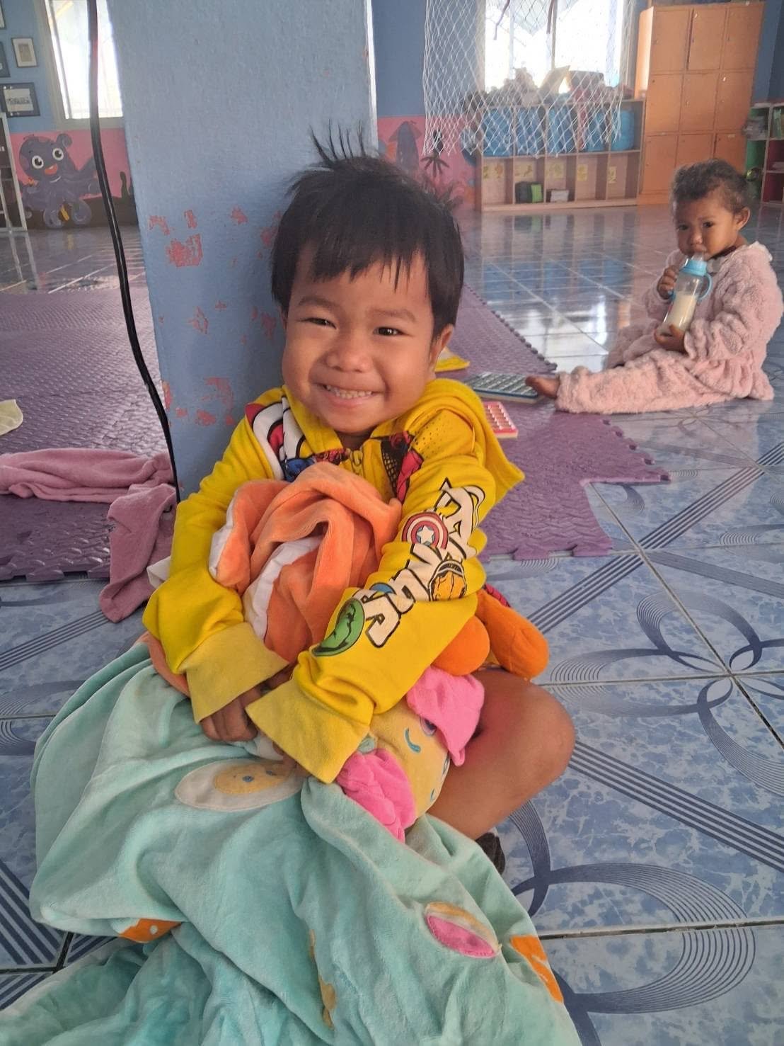 A smiling child holding a blanket in the community center, with another child in the background.